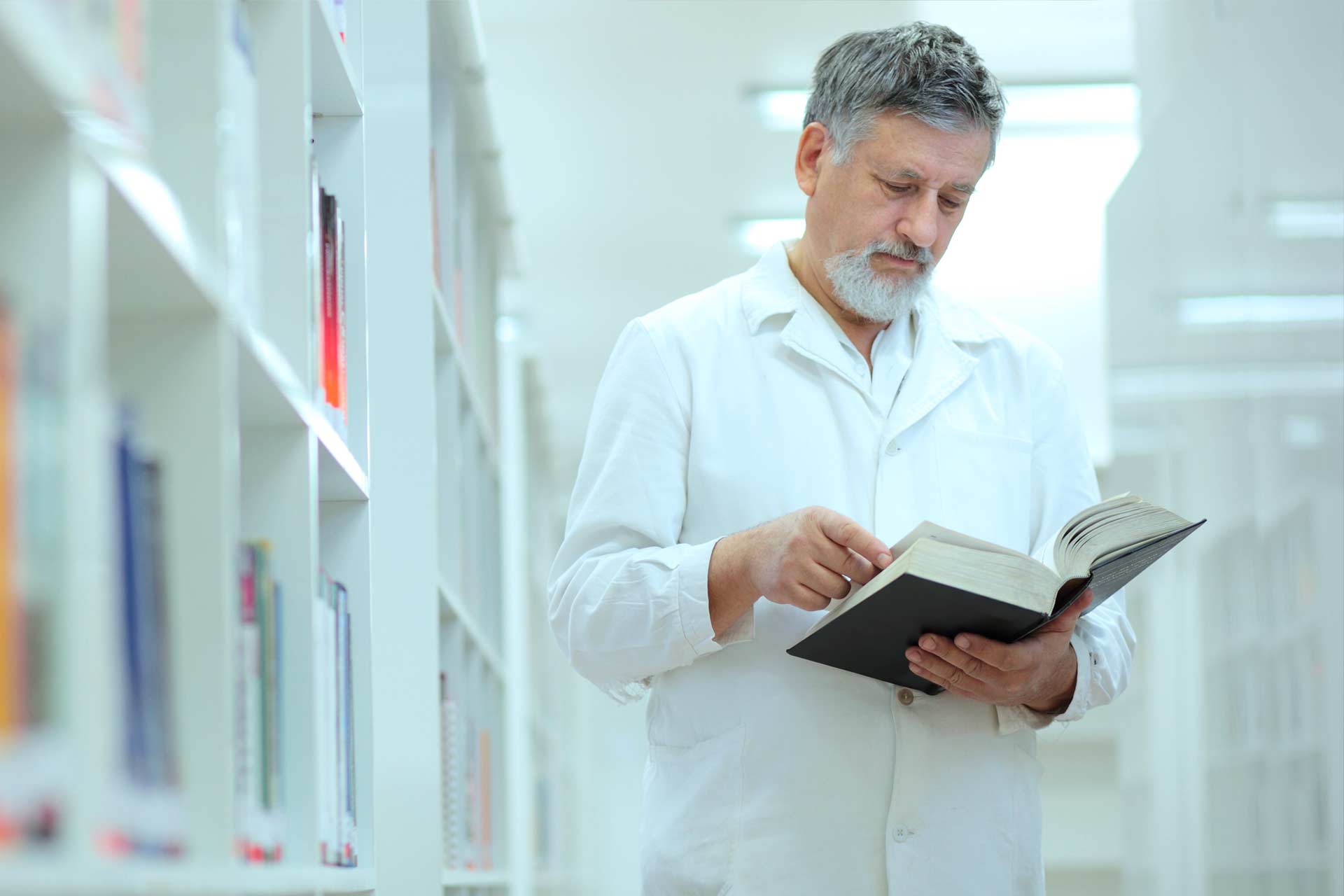 Male clinician in white coat reviewing cannabis research, standing in medical library holding thick book.