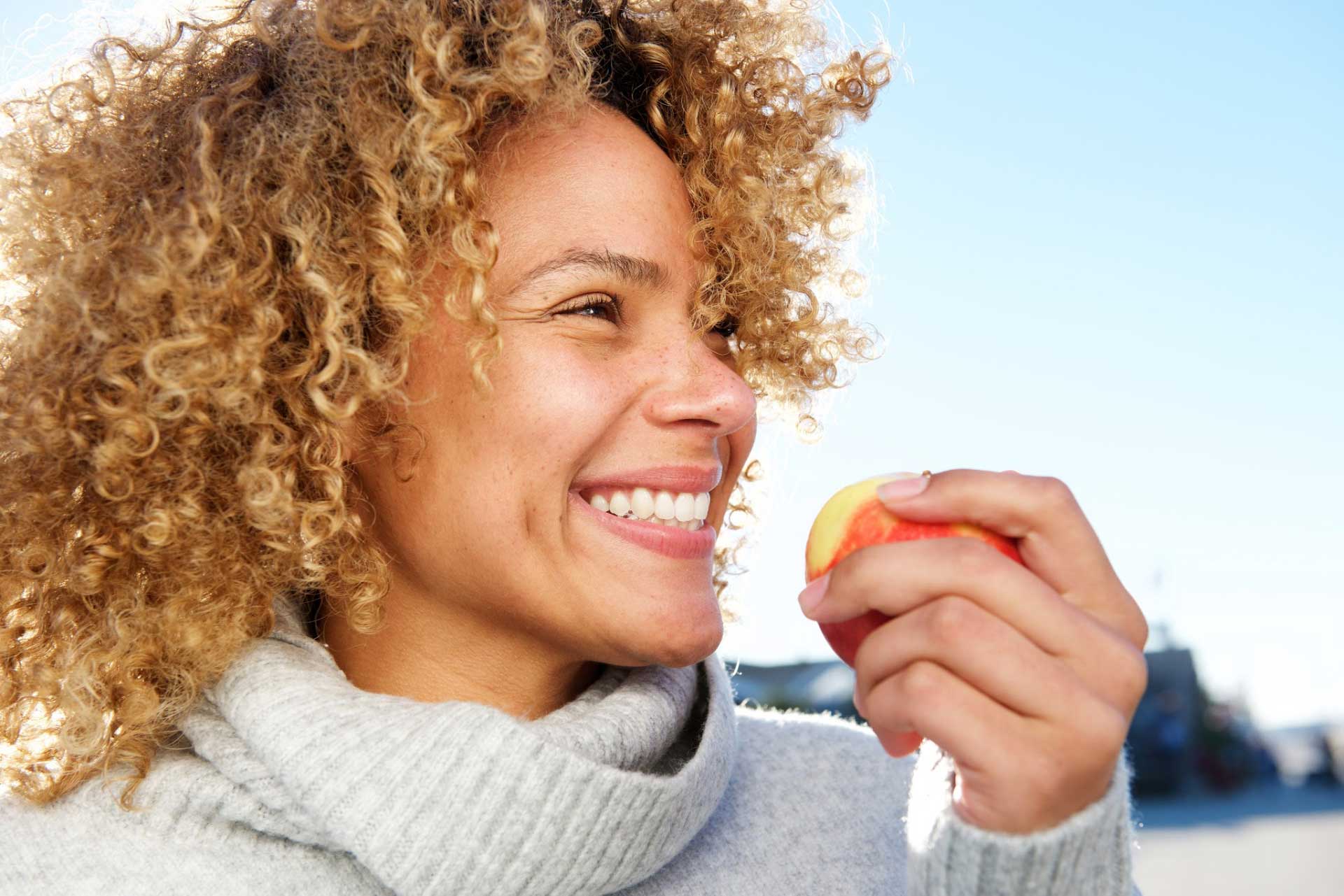 A close-up of a smiling woman standing outdoors with a fresh apple in her hand.