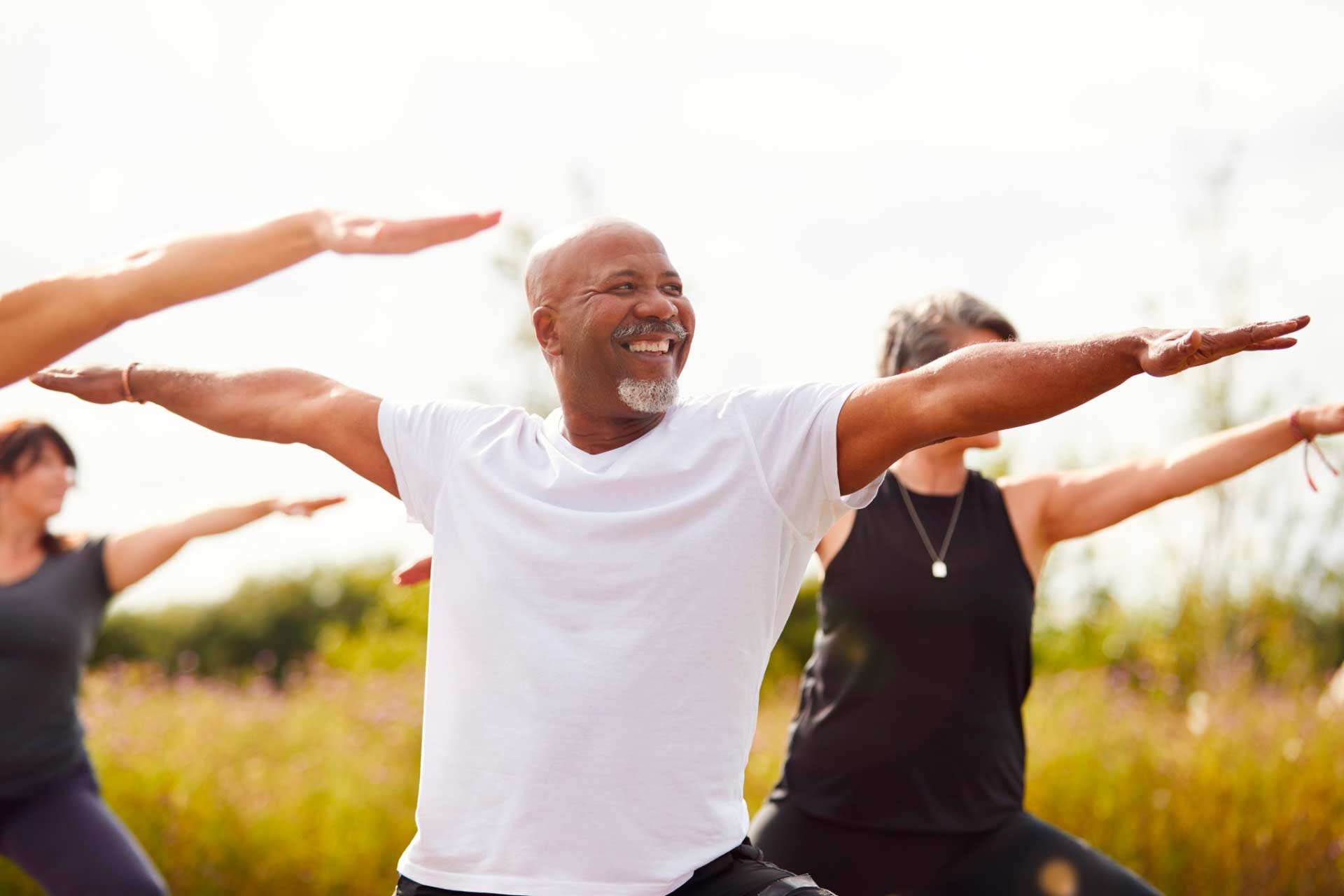 Smiling older African-American man in white t-shirt doing outdoor yoga on a sunny day, representing wellness and health.