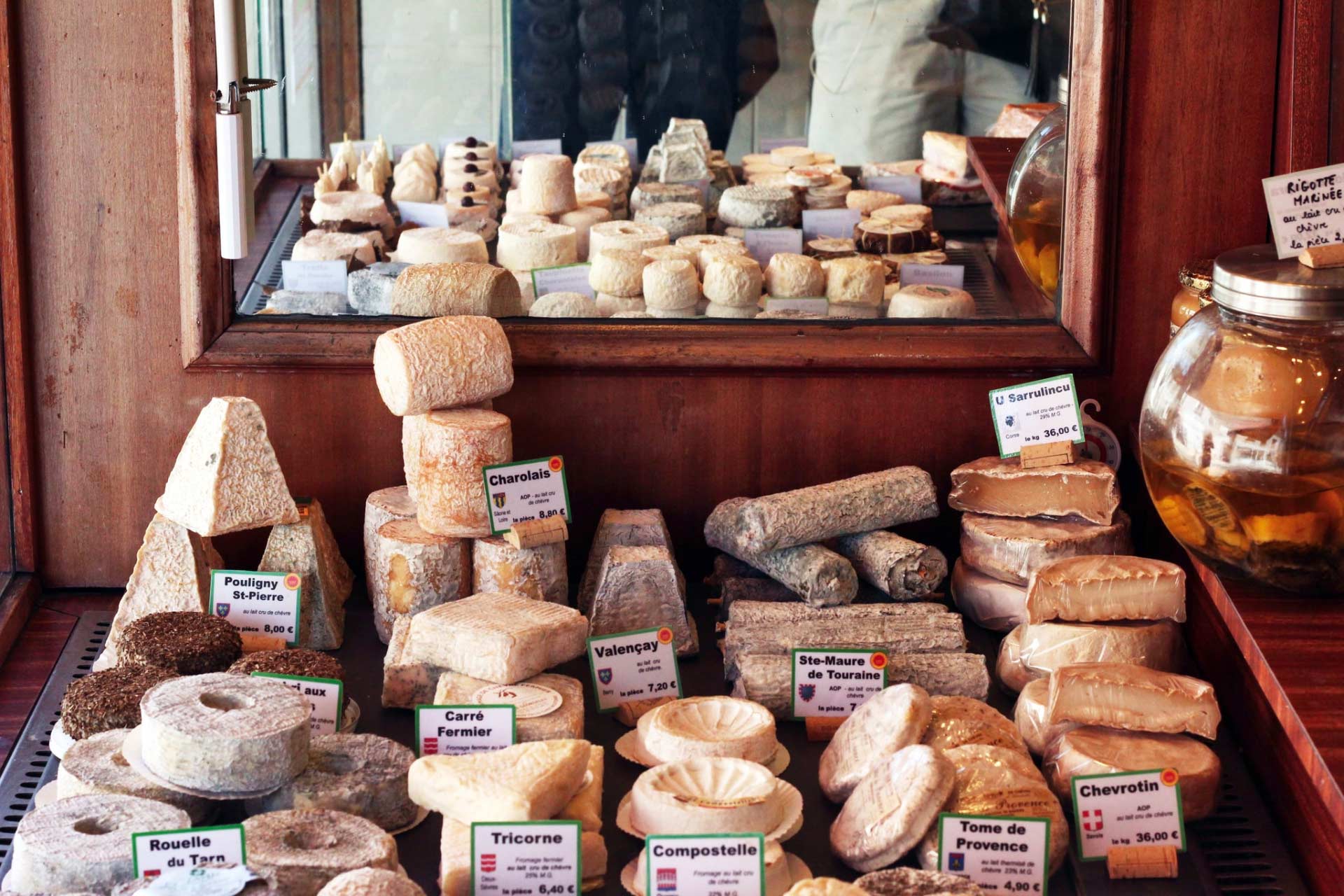 Peering inside a well-stocked cheese shop with various cheese wheels and signs showing cheese name and price.