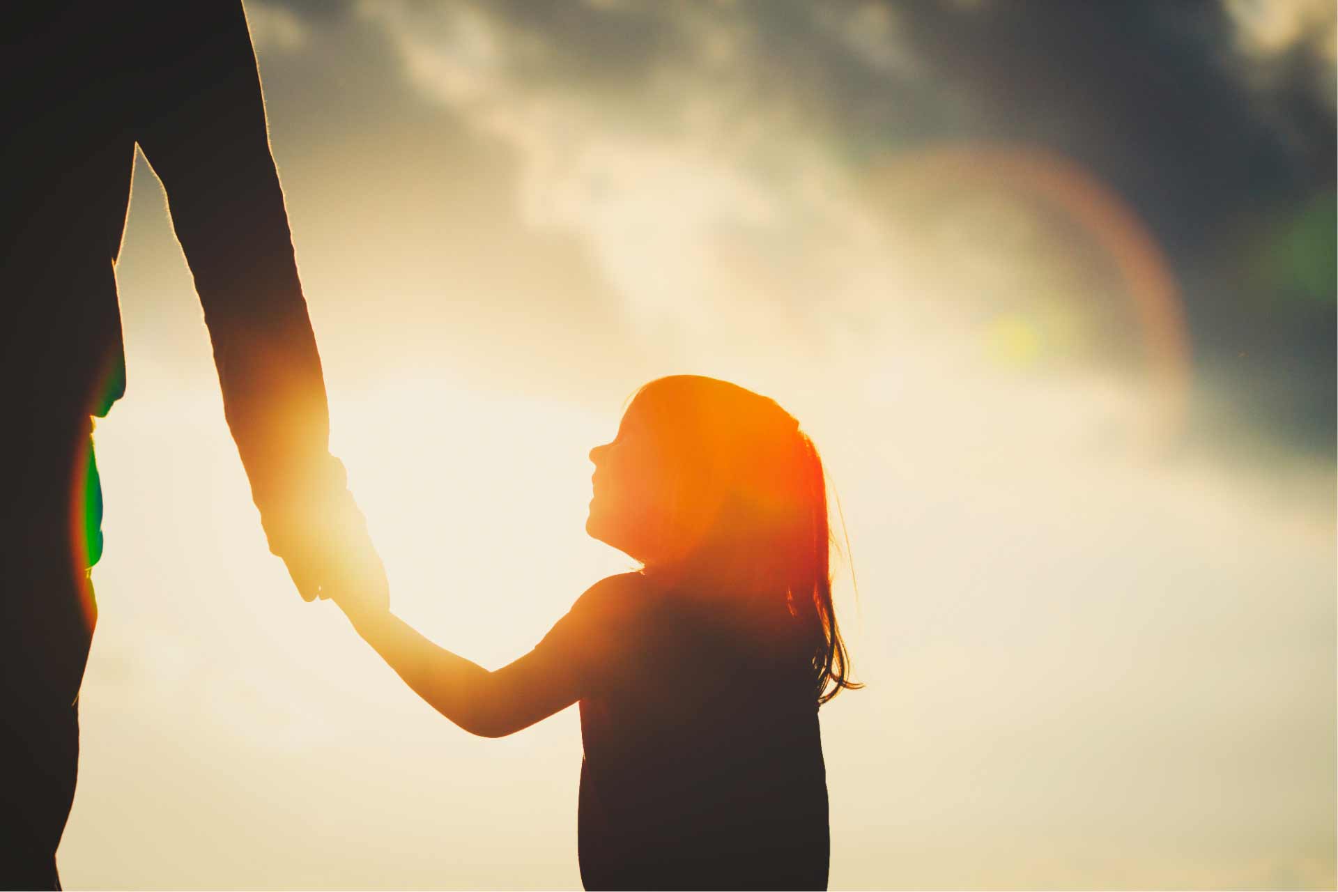 Silhouette of young girl and caregiver holding hands with sun shining in background.
