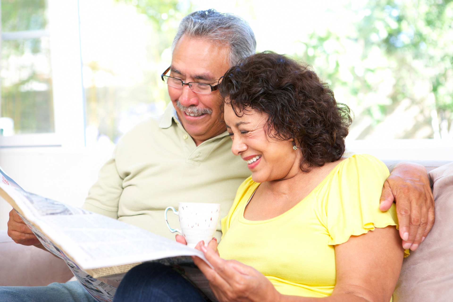 Smiling older Latino couple sits together on a sunny afternoon, reading the newspaper.