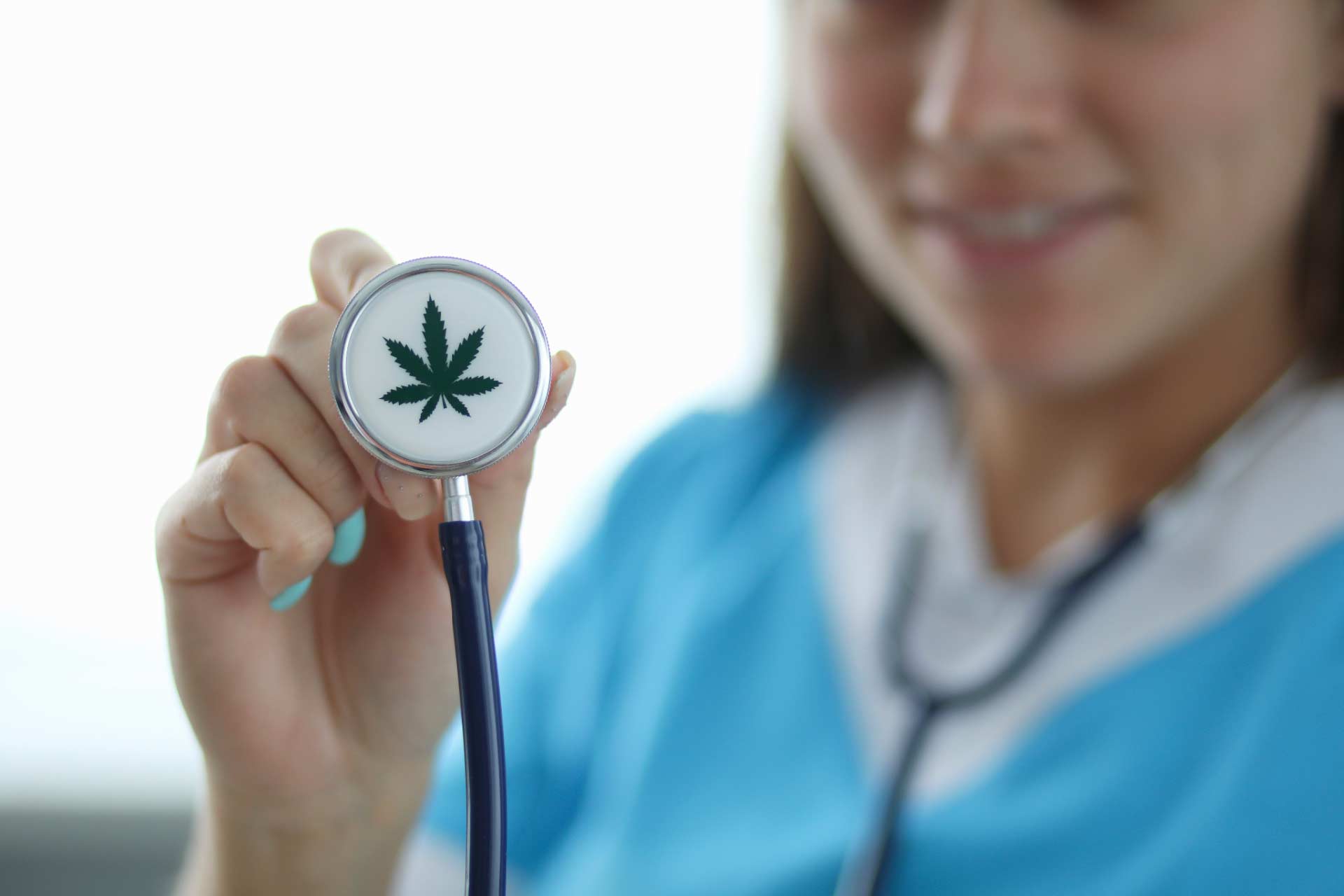 Smiling cannabis nurse in background, holding up stethoscope with a marijuana leaf image printed on it.