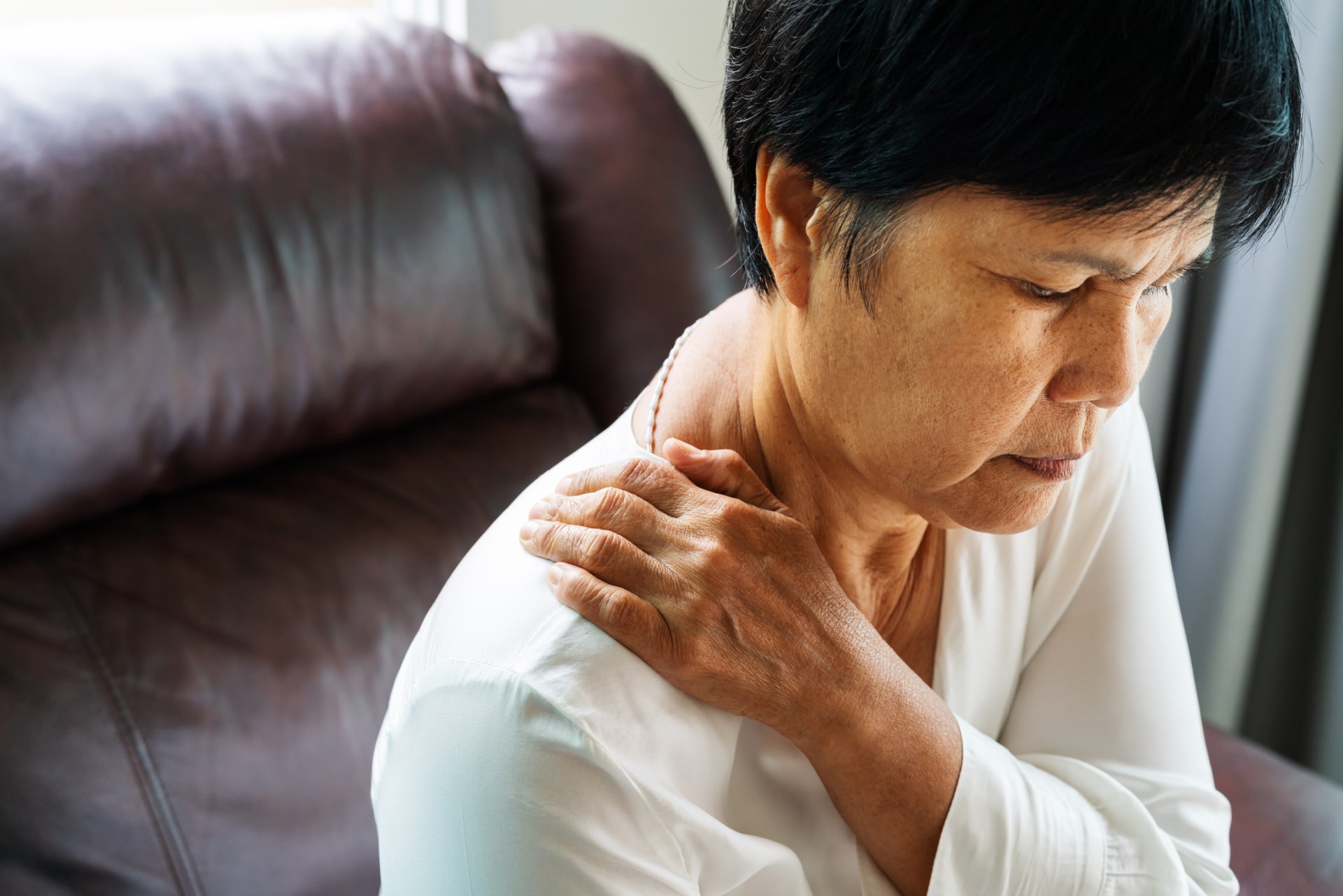 Older Asian woman holding her hand on her shoulder with expression indicating pain.