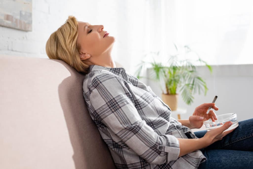side view of mature woman with closed eyes sitting on sofa and holding joint with legal marijuana