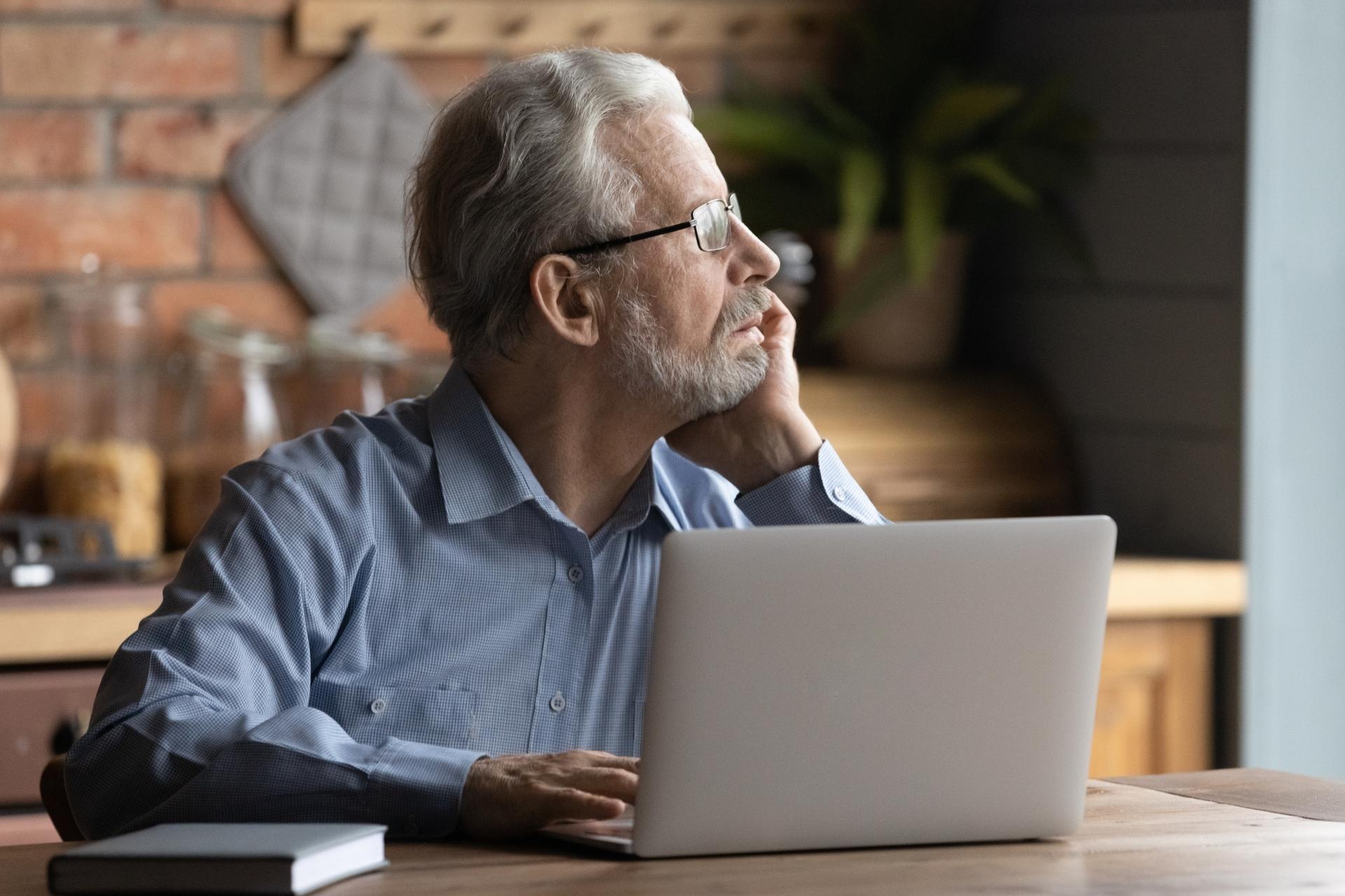 Older wellness-focused white man sitting at laptop in his kitchen, staring out window as he thinks about his CBD questions for Leaf411.