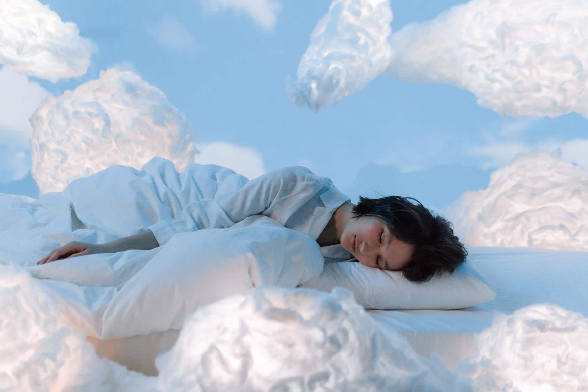 Woman with dark hair lying on her side, sleeping in the clouds with white pillows and a white blanket in a blue sky