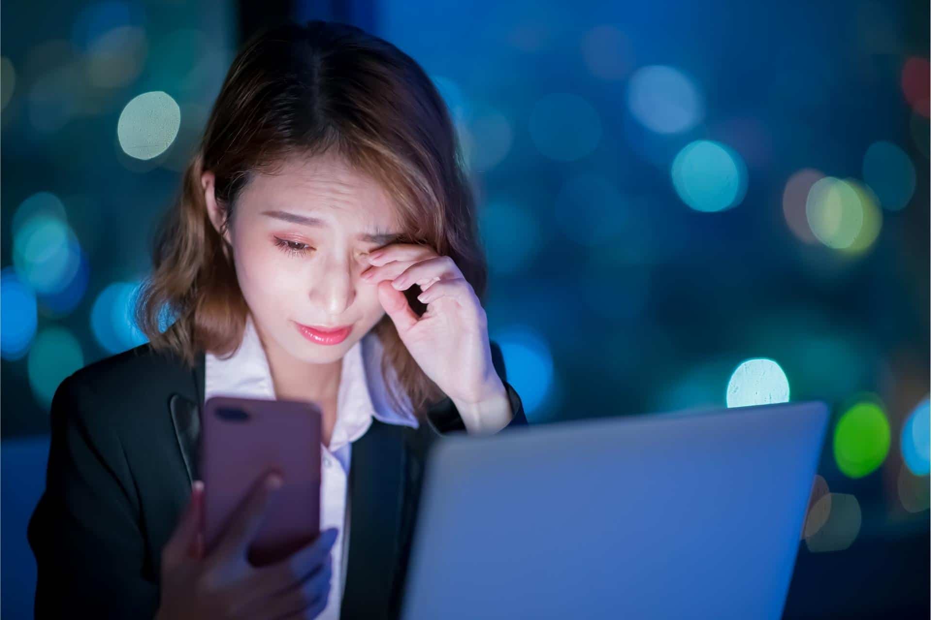 Professional woman using both laptop and smartphone at night, which may contribute to poor sleep.