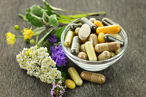 Handful of pills and supplements sitting in a clear glass bowl with plants laying around it
