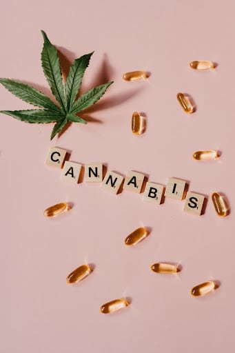 Cannabis oil capsules next to cannabis leaf on pink background with the word cannabis spelled in scrabble letters