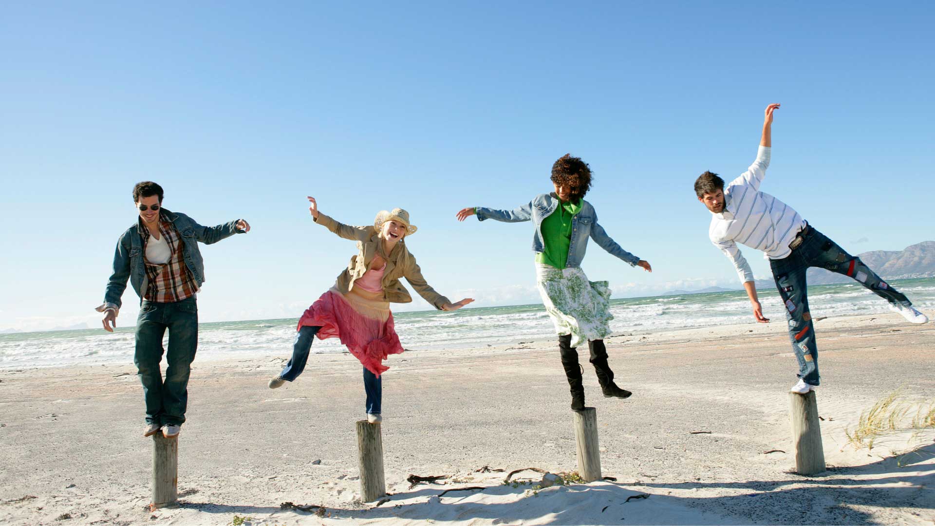 Four diverse young adults on a sandy beach, smiling while balancing on logs, representing the dynamic balance of the endocannabinoid system.
