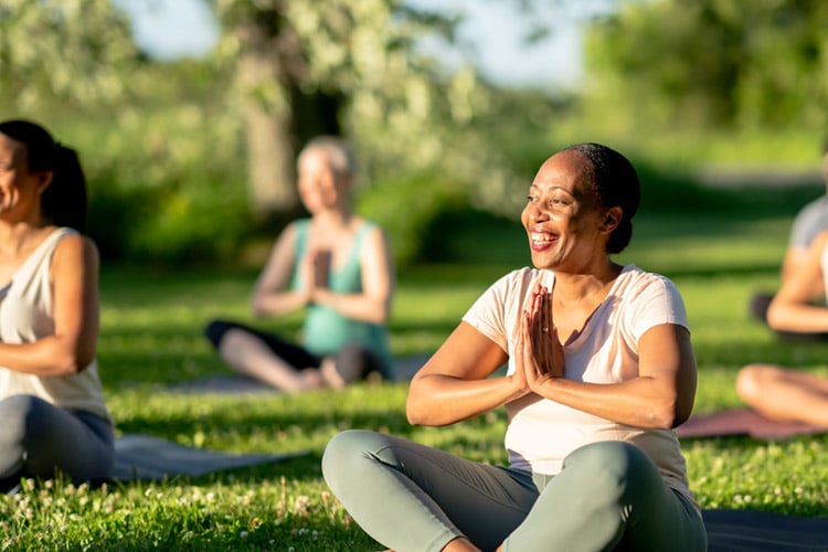 Everyone’s ECS is different in this diverse group of women doing yoga in the park, with an older smiling Black woman in the foreground.